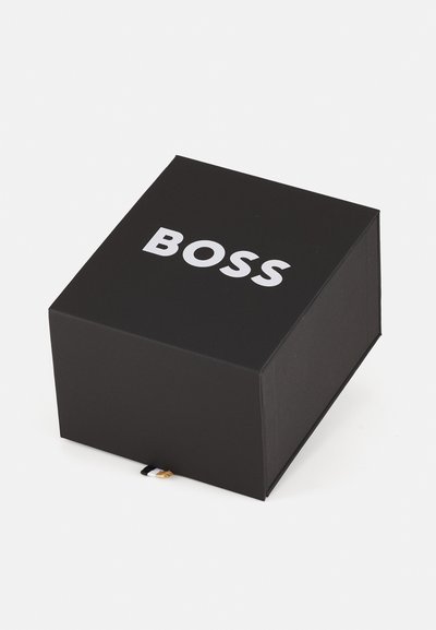 Черна правоъгълна кутия с "BOSS" с дебели бели букви отпред. Гладка матова текстура, с малък черно-бял детайл на етикет.