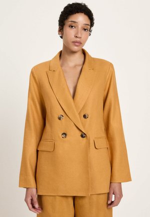 Frau mit kurzen lockigen Haaren, die einen senffarbenen, doppelt geknöpften Blazer mit Schildpattknöpfen und passende Hosen trägt, neutraler Hintergrund.