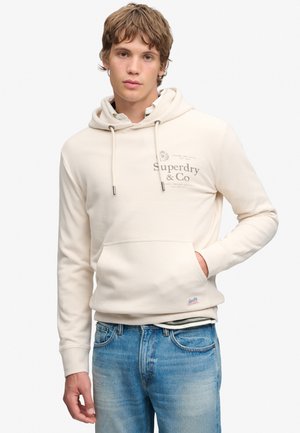 Młody mężczyzna z jasnobrązowymi włosami w kremowej bluzie z kapturem Superdry i niebieskich dżinsach, stojący z jedną ręką w kieszeni.
