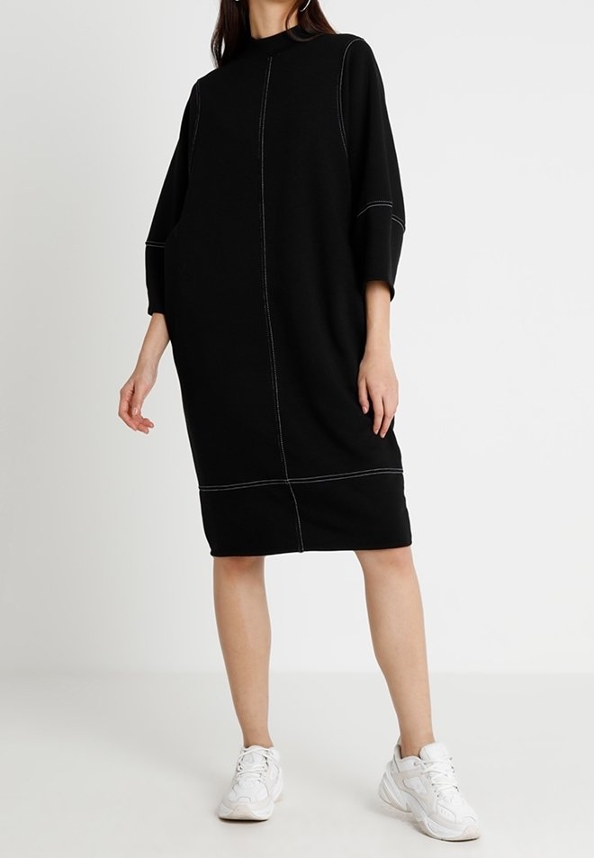 Femme portant une robe noire ample mi-longue avec des surpiqûres blanches en contraste et des baskets blanches, debout devant un fond clair uni.