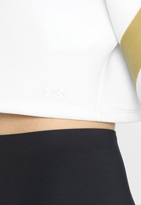 Gros plan d'une personne portant un haut blanc avec le logo Under Armour et un bas noir taille haute, montrant une partie du ventre.