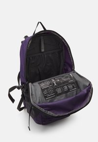 Purple and black backpack with a gray interior, featuring multiple compartments and instructional labels on a flat panel inside.