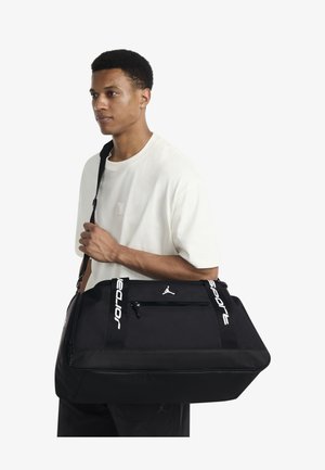 Melna somas soma ar gludu tekstūru, uz kuras redzams balts logotips un uzraksts "Sport". Tai ir rāvējslēdzēja kabata un regulējams plecu siksna.