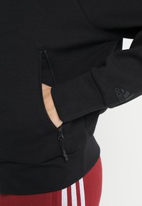 Sudadera negra con un bolsillo con cremallera en la parte frontal y puños acanalados; presenta un logo de adidas en la manga.