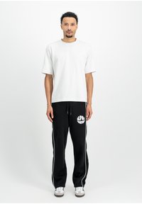 Weißes T-Shirt mit kurzen Ärmeln, kombiniert mit schwarzen Sweatpants, die ein weißes Logo und seitliche Streifen aufweisen. Graue und weiße Sneaker runden das Outfit ab.