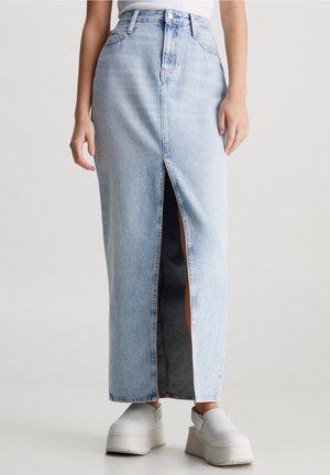 Jupe maxi en jean bleu clair avec une fente devant, portée avec des chaussures à plateforme blanches, montrée sur une personne debout devant un fond uni.