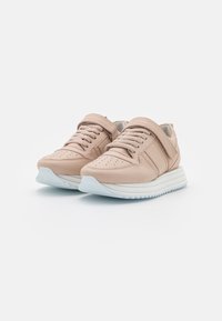 Beige sneakers dengan sol putih tebal, tali sepatu, dan strap Velcro di atas latar belakang putih.