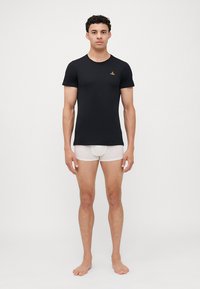 Camiseta preta de manga curta com decote redondo. Feita de um tecido suave. O modelo usa cuecas brancas por baixo. Design minimalista com um detalhe de logotipo.