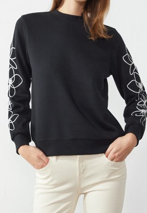 Femme portant un pull noir avec des contours floraux blancs sur les manches et un pantalon beige clair, debout les mains dans les poches.