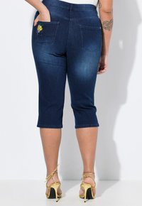 Tmavé denim capri džíny s přiléhavým střihem, ozdobené vyšitými detaily na zadních kapsách a zkráceným lemem.