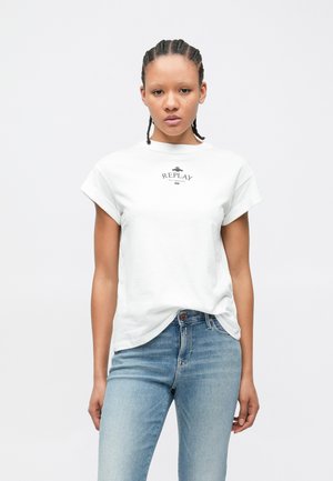 Γυναίκα με πλεξούδες στέκεται προς τα εμπρός φορώντας λευκό T-shirt Replay και ανοιχτόχρωμο τζιν μπροστά σε απλό φόντο.