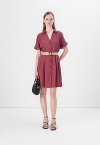Robe chemise bordeaux à manches courtes, fermeture à boutons et taille ceinturée. Les accessoires incluent un sac à main noir texturé et des sandales à lanières.