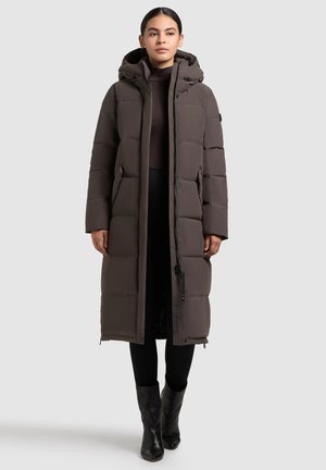Cappotto imbottito lungo con cappuccio di colore marrone scuro, caratterizzato da sezioni trapuntate, tasche con zip e zip frontale. Indossato sopra leggings neri e stivali.