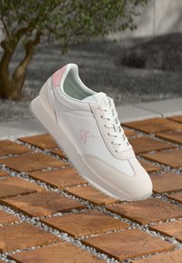 Sneaker bianco con dettagli e lacci rosa, logo "cK" di lato, sospeso su un pavimento esterno in pietra con motivo a piastrelle in legno.