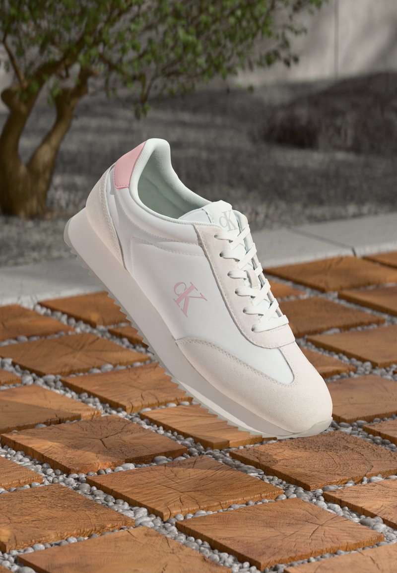 Sneaker bianco con dettagli e lacci rosa, logo "cK" di lato, sospeso su un pavimento esterno in pietra con motivo a piastrelle in legno.