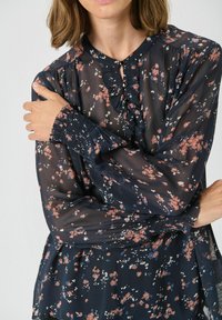 Bluză florală bleumarin din material transparent, cu mâneci lungi, manșete pliate, guler rotund și detalii cu nasturi mici în față.