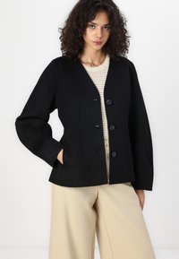 Svart, langermet jakke med V-hals, festet med tre svarte knapper, med smal midje og romslige ermer. Båret over en beige topp.