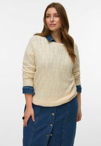 Suéter de punto trenzado en color crema con un escote amplio y puños enrollados, superpuesto sobre una camisa de mezclilla con detalles de botones, combinado con una falda de mezclilla.