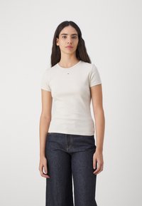 Beige ribbestr T-skjorte med rund hals og korte ermer, med en liten logo på brystet, kombinert med mørkeblå vide jeans.