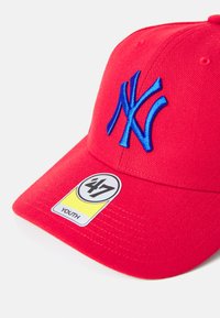 Röd basebollkeps med en stor blå broderad "NY"-logotyp, med en gul etikett märkt "47 YOUTH" på skärmen.