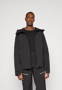 Zwarte windbreakerjas met een capuchon, voorzien van met rits gesloten zakken, een rechte snit en een matte finish. De stof lijkt lichtgewicht en gestructureerd.