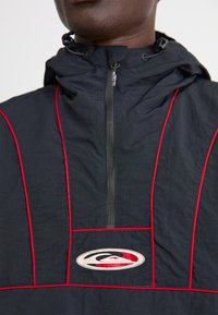 Chaqueta negra con cremallera y detalles rojos, que presenta un cuello alto, capucha y un parche con el logo en la parte frontal. El material parece ligero y texturizado.