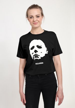Jeune femme portant un t-shirt noir d'Halloween montrant un visage masqué blanc et un texte, debout sur un fond clair uni.