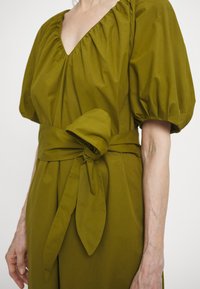 Olivově zelené šaty s výstřihem do V, nafouknutými rukávy a zavazovacími pásky v pase. Vyrobené z texturované tkaniny, s drapériemi a jemnými záhyby.