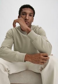 Hellbeige Sweatshirt mit gerippten Bündchen, kombiniert mit hellen Hosen, getragen von einer Person, die vor einem neutralen Hintergrund sitzt.