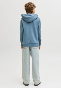 Άτομο που στέκεται με την πλάτη γυρισμένη, φορώντας μπλε φούτερ με κουκούλα, φαρδιά ανοιχτόχρωμα μπλε τζιν και μαύρα και λευκά αθλητικά παπούτσια σε απλό φόντο.