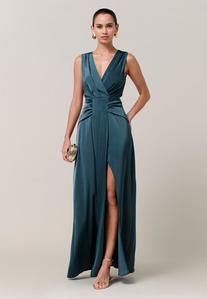 Vrouw die een teal mouwloze jurk draagt met een diepe V-hals, een hoge split tot op de dij, gouden oorringen, een gouden clutch vasthoudt en gouden sandalen met bandjes draagt.