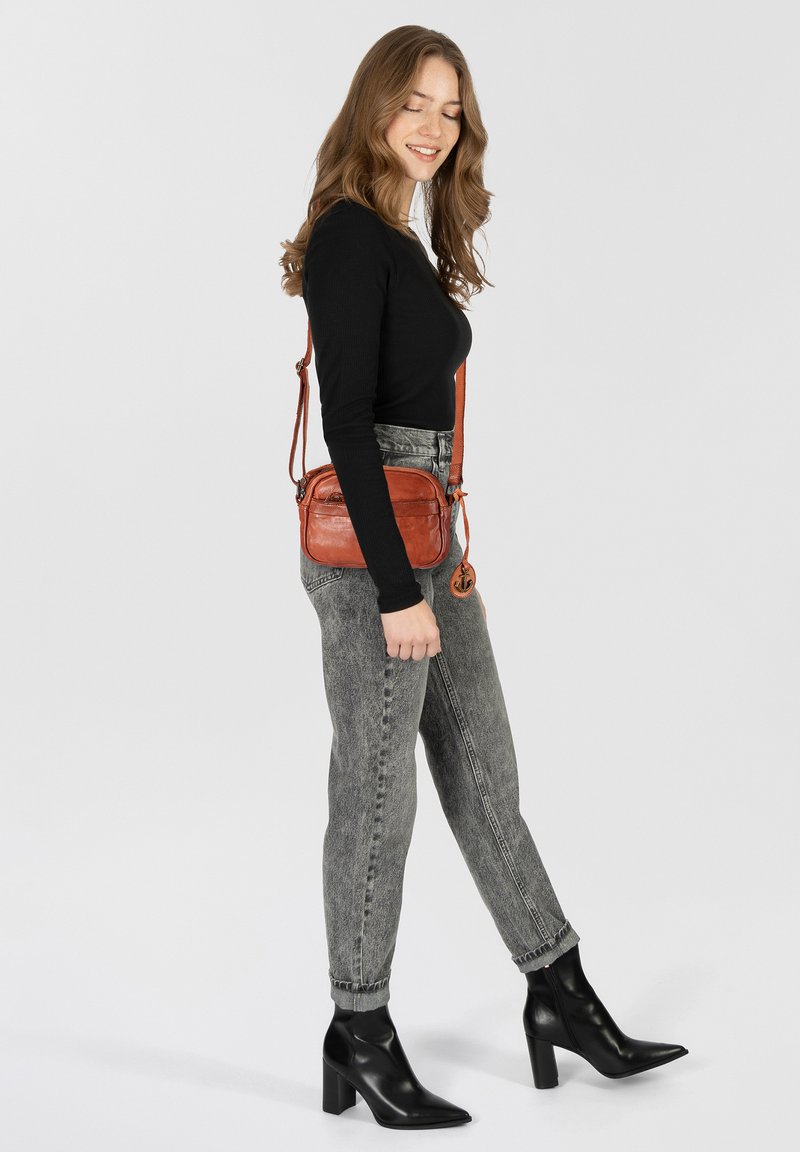 Leather crossbody bag in rust color with a textured finish, zip closure, and a detachable strap. Paired with gray denim jeans and black boots.