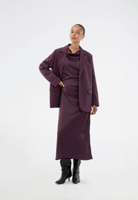 Μπλέιζερ βεραγκού σε απόχρωση δαμάσκηνου πάνω από ένα ταιριαστό σατέν φόρεμα, συμπληρωμένο από μαύρες μπότες έως το γόνατο. Λείο υλικό, κομψή γραμμή και λεπτομέρεια με μία τσέπη.