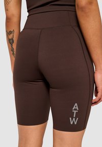 Braune Sportshorts aus dehnbarem Material, mit einem hohen Bund und kontrastierendem grauem "ATW"-Text an der Seite.