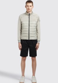 Lichtgrijze pufferjack met ritssluiting, ronde kraag en geribde manchetten, gecombineerd met zwarte shorts en witte sneakers.