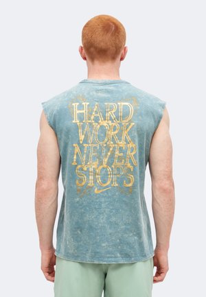 Άνδρας με κοντά κόκκινα μαλλιά φορά ένα μπλε αμάνικο πουκάμισο με την επιγραφή "HARD WORK NEVER STOPS" με μεγάλα κίτρινα γράμματα στην πλάτη.