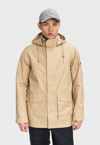 Beige wasserfeste Jacke mit Kapuze, vorderer Druckknopfverschluss, zwei großen Taschen und verstellbaren Kordeln. Graue Schirmmütze sichtbar.
