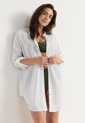 White button-up shirt made of lightweight fabric, featuring long sleeves rolled at the cuffs and a relaxed fit, layered over a green bikini.