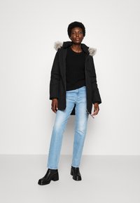 Giacca parka nera con cappuccio foderato in pelliccia, abbinata a un maglione nero a maglia e jeans azzurri chiari. Presenta stivali neri con tacco grosso.