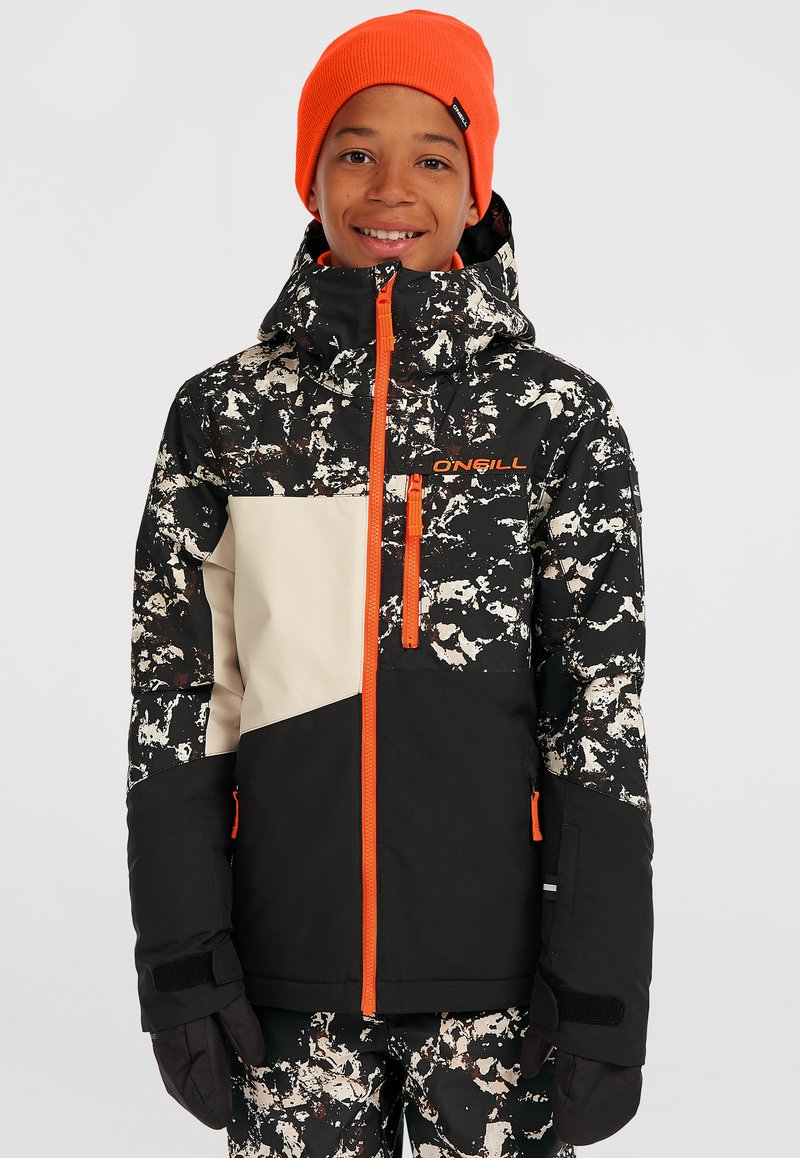 Schwarz-weißer gemusterter Winterjacke mit orangefarbenen Akzenten, einem hohen Kragen und mit Reißverschlüssen versehenen Taschen; getragen mit einer orangefarbenen Strickmütze.