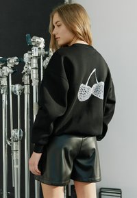 Svart oversized sweatshirt med vitt virkade körsbärsdesign på ryggen, matchat med blanka svarta läderbyxor. Studio bakgrund och belysningsutrustning.