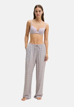 Femme debout pieds nus portant un soutien-gorge violet clair et un pantalon de pyjama ample à carreaux, les mains dans les poches, sur un fond uni.