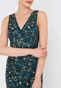 Green sequined dress with a V-neck and floral embroidery in pink, blue, and gold. Features textured fabric with intricate detailing.