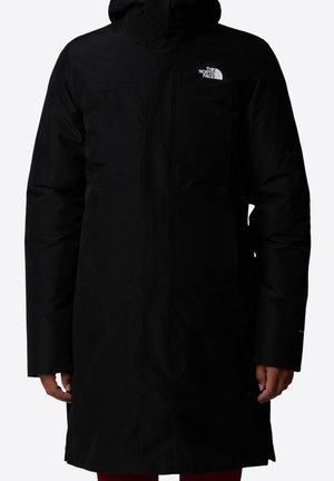 Black long winter jacket with hood, front zipper, and white The North Face logo on the chest, worn by a person with hands relaxed at sides.