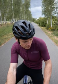Schwarzer Fahrradhelm, passendes kurzärmliges Trikot in Dunkelpurpur mit Reißverschluss, schwarze Fahrradhosen und ein Schotterweg, gesäumt von Bäumen.