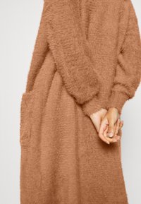 Personne portant une robe-pull camel floue à manches longues, les mains jointes dans le dos, montrant le tissu texturé et une poche latérale.