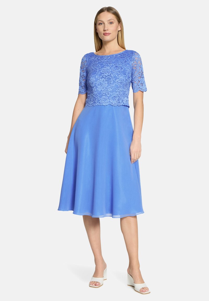Robe bleue avec un corsage en dentelle à manches courtes, jupe fluide en chiffon, et ourlet arrivant au genou. Portée avec des sandales blanches à talons blocs.