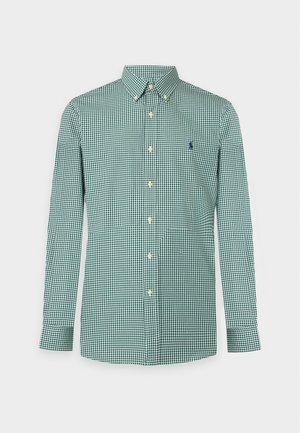 Homme portant une chemise verte à carreaux manches longues, un short beige à hauteur des genoux et des chaussures beiges sans lacets, debout avec une main dans la poche.