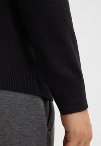 Maglione nero lavorato a maglia con polsini a costine, che presenta una texture liscia e un design aderente, indossato sopra pantaloni della tuta grigi.