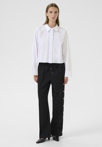 Biała krótka koszula z zapięciem na guziki z przodu, zestawiona z luźnymi czarnymi spodniami z bocznymi akcentami guzikowymi. Teksturowana tkanina.